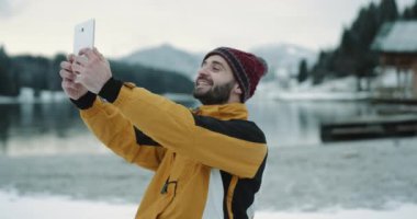 Çok komik turistik karizmatik surat selfies muhteşem manzaralı göl ve snowy orman ve dağ, bir turuncu ceket ve kırmızı şapka giyiyor turist ortasında bir tablet ile.