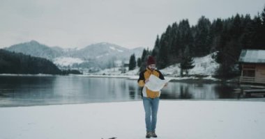 O karlı dağ ve bir ahşap evin yanında orman ile inanılmaz manzara mükemmel kış günde geldi bir harita ile karlı bir göl çevresinde yürüyüş harita ile karizmatik genç turist. 4k