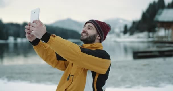 非常有趣的旅游魅力的脸, 使自拍与平板电脑在中间的景观惊人的看法湖和雪林和山, 游客穿着橙色夹克和红色帽子. — 图库视频影像