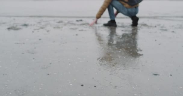 Ein fröhlicher junger Mann mit einer blauen Tasche auf dem Rücken verbringt viel Zeit am Strand neben dem blauen Meer, nimmt nassen Sand in die Hand und spielt. — Stockvideo