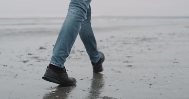 L'heure de la plage sur un homme de jour de pluie marchant à travers le bord de la mer, capturant la vidéo de l'arrière des jambes des hommes. 4k — Video