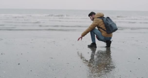 Jongeman die het grijze strand verkent na een regenbui aan de kust, filmt van dichtbij, neemt wat nat zand en gooit het water in. 4k — Stockvideo