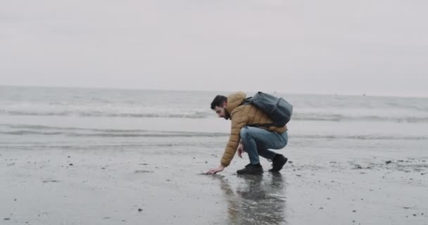 Giovane felice di esplorare la spiaggia dopo una pioggia lungo la riva del mare è geo molto grande e di buon umore, prendere un po 'di sabbia bagnata e buttare giù. 4k — Video Stock