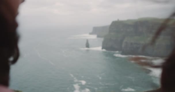 Due giovani signore sulla cima delle scogliere con le spalle alla macchina fotografica che mostrano un grande cuore con le mani che guardano verso l'oceano, in una giornata ventosa. 4k. rallentamenti . — Video Stock