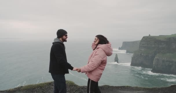 Jong koppel gelukkig reizen samen staan zij op de top van Cliffs of moher en onder de indruk op zoek naar het uitzicht op de Oceaan, man aan haar en maken een voorstel op de top van Cliffs dame erg blij en — Stockvideo