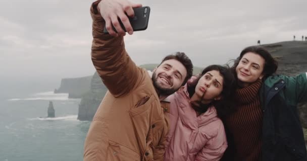 Красивые друзья мульти этнического фотографирования на вершине скалы Мохер с помощью смартфона, они делают смешные лица и улыбаются большие, принимая видео крупным планом . — стоковое видео