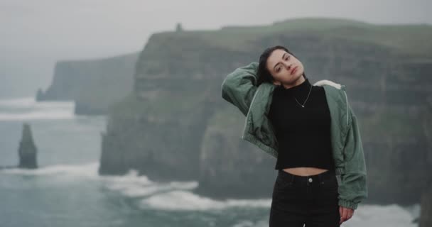 Linda senhora na frente da câmera em pé na margem de falésias de Moher e carismático olhando diretamente para a câmera. movimentos lentos — Vídeo de Stock