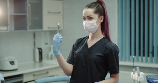 Em uma sala de clínica moderna, uma jovem dentista preparando a seringa para fazer uma injeção, olhando diretamente para a câmera. 4k — Vídeo de Stock