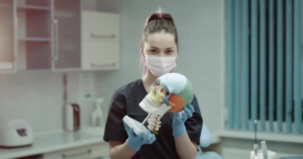 Jeune assistante médicale portant un masque chirurgical et des gants et devant la caméra dans une salle de clinique moderne examinant un crâne humain . — Video