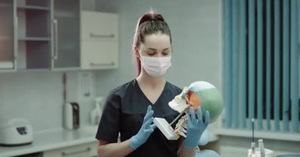 Retrato de una joven doctora con una máscara quirúrgica y guantes en la moderna sala de la clínica sosteniendo un gran cráneo humano y mostrando a la cámara. 4k — Vídeos de Stock
