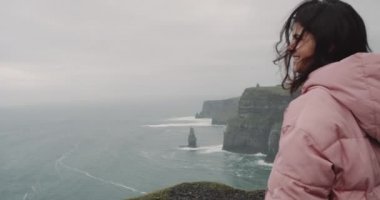 Genç kadın o sahile duran geldi uçurumun tepesinde şaşırtıcı yerde ve manzara okyanus manzarası ve kayalıklarla hayran. 4k.