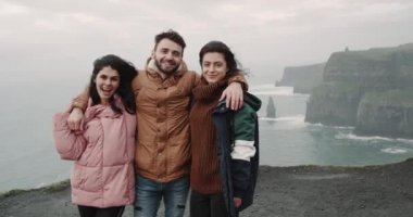 Moher kayalıklarla muhteşem okyanus manzarasına sahip muhteşem yerde anılar fotoğraf çekmek için kamera önünde poz mutlu üç arkadaş turist. 4k