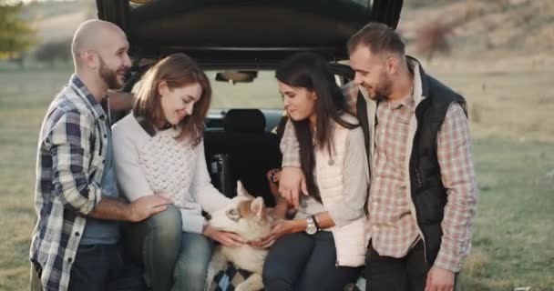 Merveilleux deux couples assis sur le coffre de la voiture et passer un moment de détente en souriant et en bavardant ensemble par une journée ensoleillée, au milieu sont incroyables petit chien husky . — Video