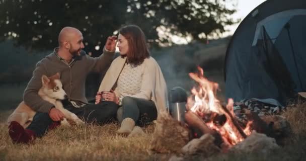 Charismatisches junges Paar verbringt eine romantische Zeit inmitten der Natur am Abend neben dem Lagerfeuer und entspannen mit ihrem Husky-Hund. — Stockvideo