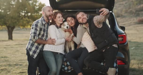 Pareja carismática en un viaje en coche tienen una parada para tomar algunas fotos selfie con el teléfono que se sientan en un maletero del coche . — Vídeos de Stock