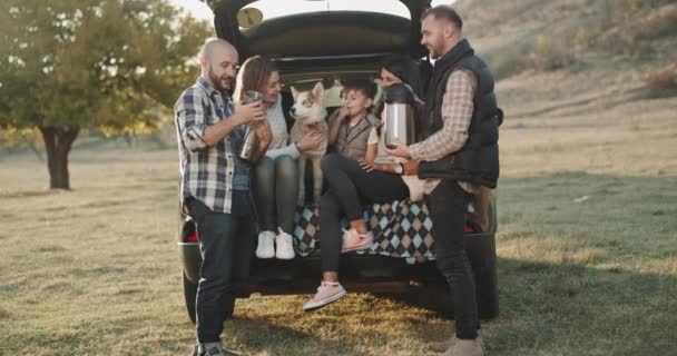 Picknickzeit für zwei Familien, die auf der Rückbank des Autos auf dem Kofferraum sitzen und glücklich Tee aus einer großen Thermoskanne trinken, kleiner Junge und ein Husky-Hund sitzen neben. — Stockvideo