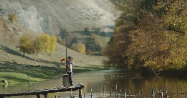 Um menino bonito segurando uma grande vara de pesca em uma ponte ao lado do lago e jogando . — Vídeo de Stock