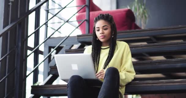 Afrikanerin mit langen afrikanischen Haaren sitzt auf der Treppe und arbeitet glücklich an ihrem Notizbuch, sie lächelt und verbringt eine schöne Zeit. — Stockvideo