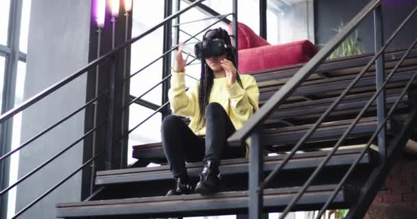 Good looking african lady using virtual reality glasses and enjoying the time using them in a modern building, she sitting down on the stairs beside of a big window. — Stock Video