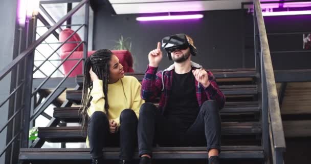 Красива африканська леді і харизматичний хлопець, граючи з окулярами віртуальної реальності і насолоджуючись разом в сучасному бізнес-центрі, вони сидять на сходах. 4k — стокове відео