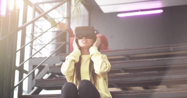 Emocionada mujer africana descubre las gafas de realidad virtual que está sentada en las escaleras y vistiendo la VR feliz empezando a explorarlas. 4k movimientos lentos — Vídeo de stock