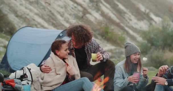 Una joven pareja y sus amigos pasando un buen rato juntos en el camping, por la noche alrededor de la fogata comer malvaviscos y beber vino . — Vídeos de Stock