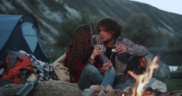 Hermosa captura de una pareja que pasa una velada romántica en medio de la montaña junto a la fogata y bebiendo algunas bebidas calientes y comiendo malvaviscos . — Vídeo de stock