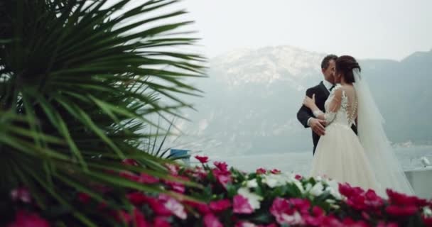 Dacing bruden och brudgummen i fantastisk plats mitt i bergen med sjön i bakgrunden, multicolor blommor och gröna blad. — Stockvideo