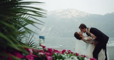 Romantik ve güzel bir çift damat ve Gelin çiçek şaşırtıcı yerde onlar tutarak ve birbirlerini öpmeye alan arka plan dağ ve göl. 4k