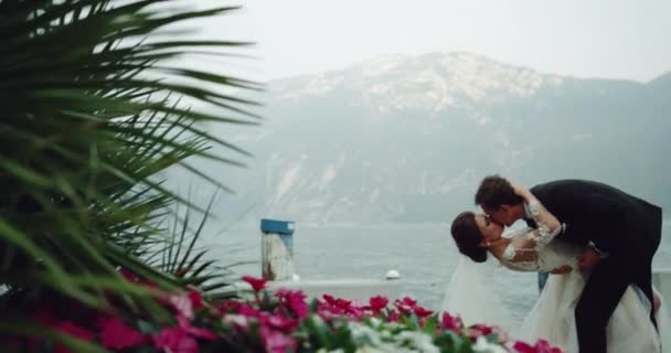 Καταπληκτικό νύφη σε λευκό μακρύ φόρεμα φιλιά ρομαντικό με το γαμπρό σε ένα υπέροχο τοπίο όμορφο τόπο στο παρασκήνιο λίμνη βουνό και όμορφα λουλούδια. — Αρχείο Βίντεο
