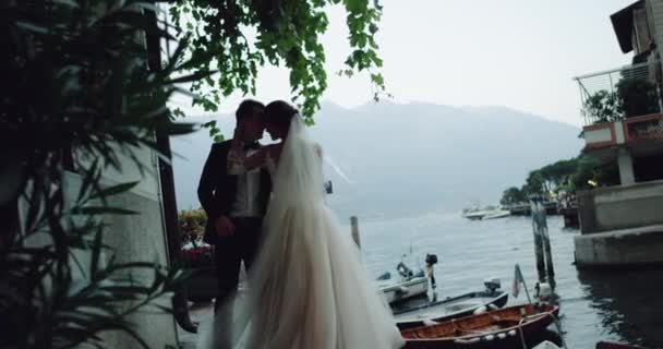 漂亮的新娘与长礼服和新郎亲吻和拥抱在惊人的地方与湖和小船旁边山在背景. — 图库视频影像