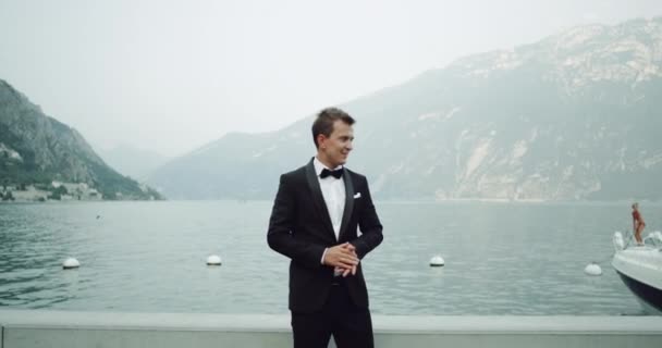 Gut aussehender junger Bräutigam im schwarzen Anzug inmitten einer atemberaubenden Landschaft mit See und Bergen, die auf die Braut warten. — Stockvideo