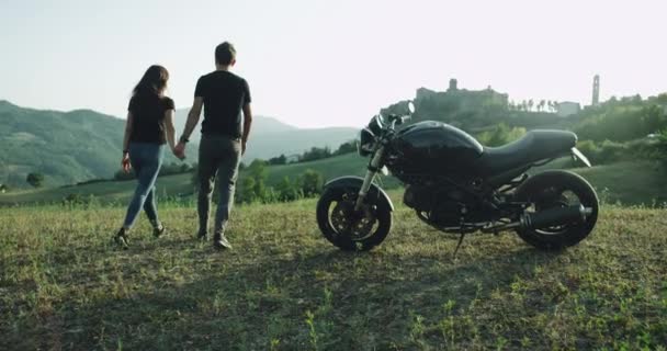 Inmitten einer grünen Wiese und wunderschönen Landschaft amüsiert sich ein Paar, sie gehen das Feld hinunter, neben ihnen steht ein großes schwarzes Fahrrad. — Stockvideo