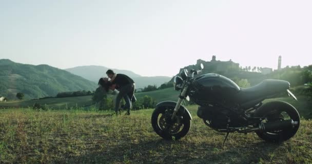 Красива пара проводить романтичний час разом у дивовижному місці в центрі природи вони обіймають один одного, поряд з ними стоїть чорний мотоцикл — стокове відео