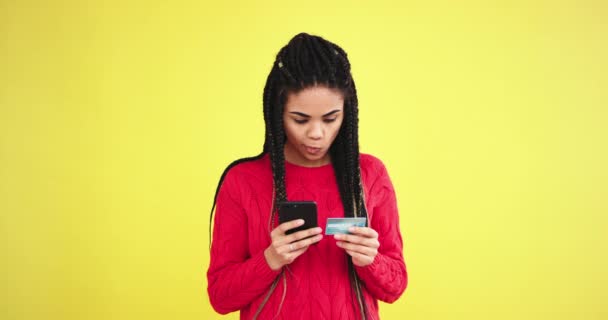 Em um estúdio com fundo amarelo parede senhora africana em uma camisola vermelha na frente da câmera usando um smartphone e cartão de banco para encomendar algo — Vídeo de Stock