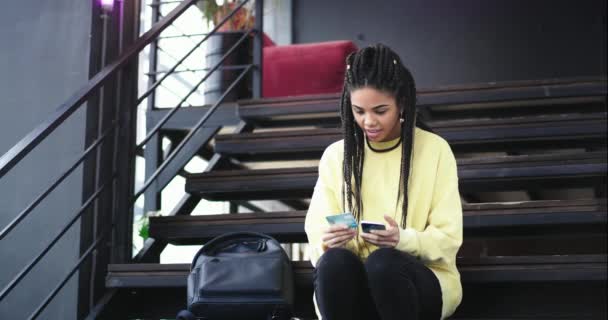Frumoasă tânără africană care își folosea smartphone-ul pentru a comanda ceva online de pe cardul ei bancar, ea fericită la sfârșit a ridicat mâinile în sus, ea stând jos pe scările moderne. mișcări lente — Videoclip de stoc