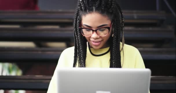 Belle femme africaine utilisant sa carte bancaire à l'aide de son ordinateur portable pour vérifier ou acheter quelque chose en ligne, elle porte des lunettes, au milieu du hall du bureau, elle s'assied sur les escaliers — Video