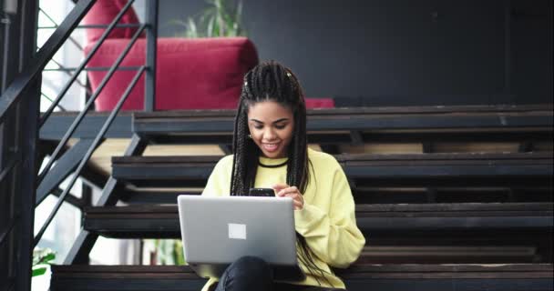 Hezká kancelář pracovník africké etnické slečna pracující v hale moderní kancelář vsedě na schodech a psát něco o její notebook velmi koncentrované — Stock video