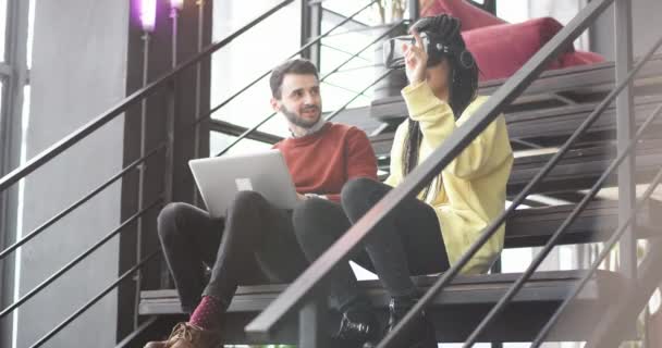 Dans un bâtiment d'affaires moderne, un gars et une dame africaine passent un bon moment ensemble assis sur l'escalier moderne dame essayant pour la première fois la VR qu'elle touche virtuel avec le doigt pour sélectionner favori — Video