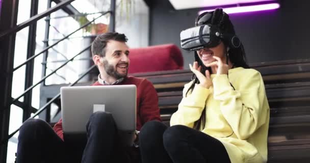 Retrato carismática mujer africana con una gran sonrisa utilizando unas gafas de realidad virtual para explorar el mundo virtual que se sienta junto a un trabajador de oficina en las escaleras, el hombre que trabaja en su computadora portátil en — Vídeos de Stock