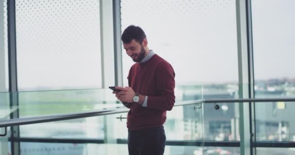 Happy business worker standing beside of a big window view and typing something in his smartphone at the same time he are smiling. 4k — Stock Video