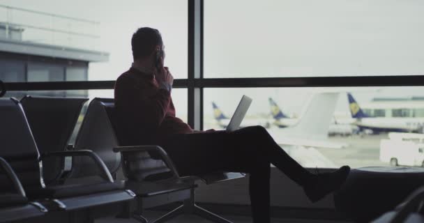 Knap jongeman in de luchthaven spreken op zijn telefoon op hetzelfde hij zijn notebook gesloten en gaan krijgen van de aanhouding — Stockvideo