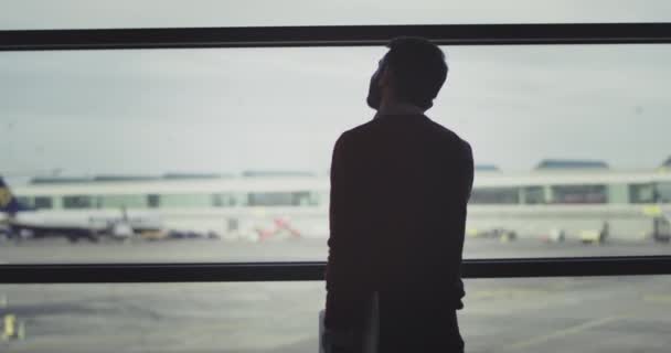 Joven hombre de negocios en el aeropuerto tienen una conversación en su teléfono hasta que espera su vuelo, que afirma con la espalda a la cámara y la celebración de su ordenador portátil — Vídeos de Stock