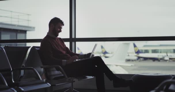 Heureux dans un homme de bonne humeur assis sur les chaises de l'aéroport attendant son vol, il utilise un ordinateur portable pour taper quelque chose puis fermé et attendant l'embarquement. 4k — Video