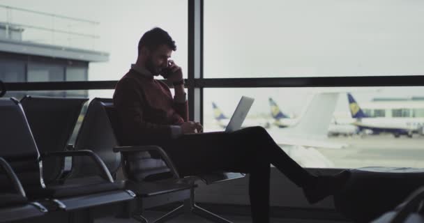 Happy with a good mood businessman working on his laptop and speaking on his phone sitting on the chair in airport, he waiting for the flight, to get boarding — стоковое видео