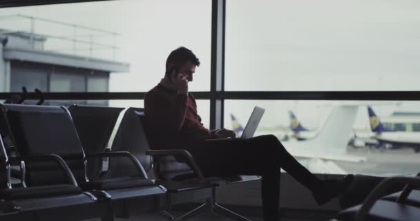 Харизматичный парень в аэропорту сидит на стуле, разговаривает по смартфону и начинает работать на ноутбуке, в то же время очень взволнованный, ждет посадки — стоковое видео