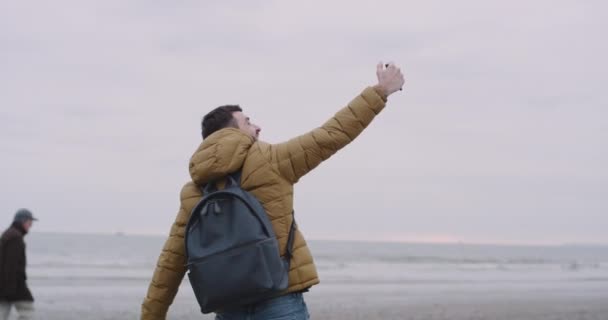 Ευτυχισμένος συνεπαρμένος τουριστικά άλματα και χαμογελώντας ευτυχισμένη σε μια μεγάλη παραλία με θέα τη θάλασσα, αυτός κρατώντας ένα τηλέφωνο και λαμβάνονται selfies της θάλασσας — Αρχείο Βίντεο