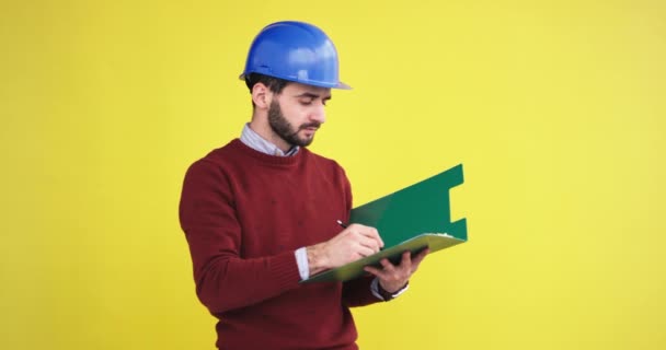 Jefe del sitio joven en el estudio con una pared de fondo amarillo con un casco azul de seguridad analizando algo y tomar las notas en el mapa de la oficina, tiene una cara concentrada . — Vídeos de Stock