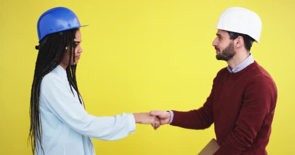 Im Atelier mit gelbem Hintergrund zwei Architekten, eine afrikanische Dame und ein Mann mit blau-weißem Helm vor der Kamera machen viel und halten am Ende Händchen — Stockvideo