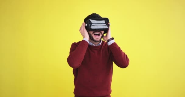 Homem vestindo um óculos de realidade virtual em um estúdio com uma parede de fundo amarelo e explorando o mundo virtual muito animado — Vídeo de Stock